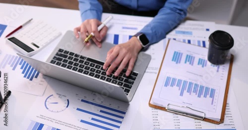 A professional businessman works diligently on his laptop at an office desk, surrounded by financial accounting documents and reports while writing notes in a notebook
