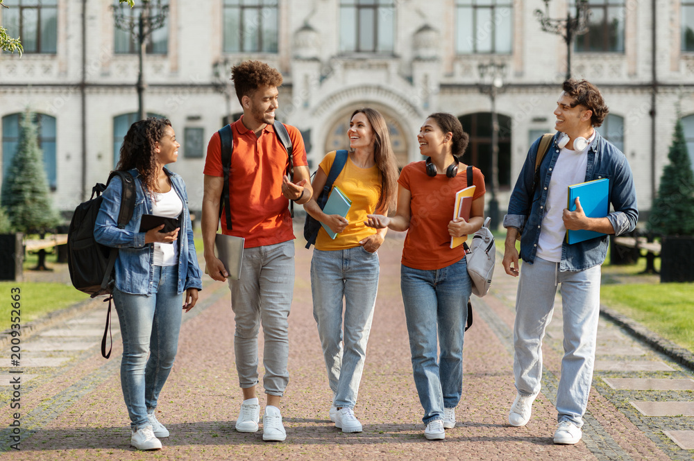 Fototapeta premium Happy university students walking together on campus, group of cheerful young multiethnic people chatting and laughing outdoors during break, carrying backpacks and workbooks, full length shot