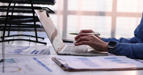 A professional businessman works diligently on his laptop at an office desk, surrounded by financial accounting documents and reports while writing notes in a notebook
