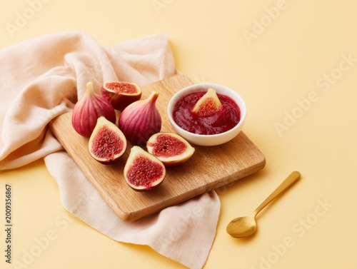 Minimal food scene with fresh figs on wooden board, red jam in white bowl, gold spoon and beige cloth on soft yellow background, clean elegant presentation.