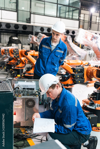 Two workers are focused on repairing machinery in a warehouse. They wear safety gear and handle tools while discussing the task at hand. The scene shows an active work environment.