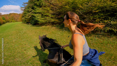 Foto SELFIE: Woman horseback riding across a sunny meadow by the lush green forest on sunny day
