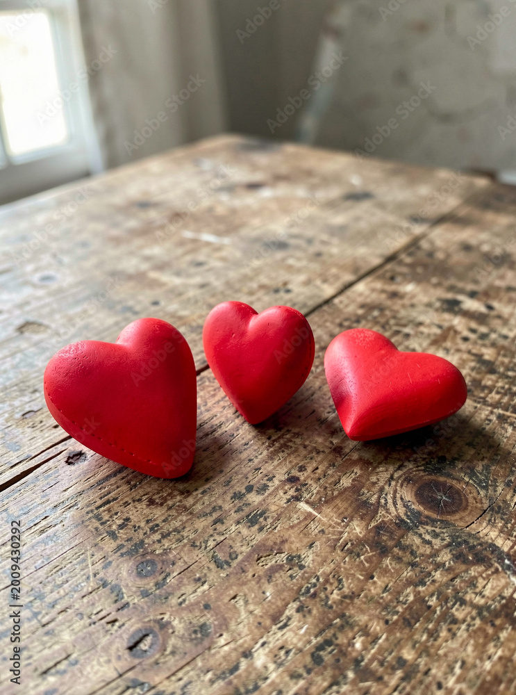 Fototapeta premium The image shows three red hearts resting on a wooden surface with visible grain and texture. The background is indistinct but appears to be an interior space.