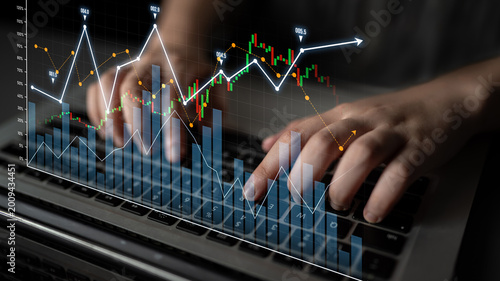 Bild auf Leinwand Close-up view of hands typing on a laptop with overlaid financial graphs and market trends, illustrating stock analysis and investment strategies in a digital workspace