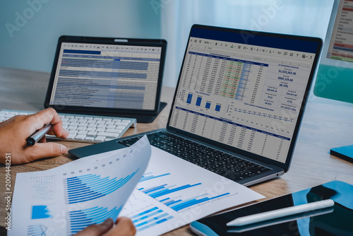 Hands reviewing financial chart document with laptop displaying spreadsheet dashboard and tablet screen, representing data analysis workflow, reporting process, business performance tracking, and digi