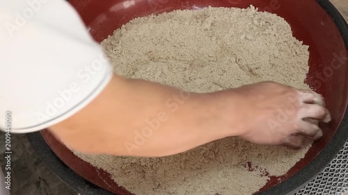 close-up shot of a person’s hand pouring water into a large red bowl filled with coarse whole wheat flour (atta). The video captures the initial step of the traditional dough-kneading process for maki