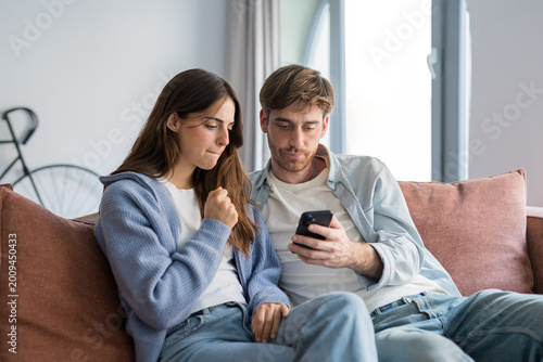 Konstfotografi A young couple sits close on a living room sofa, focused on a smartphone as they read messages