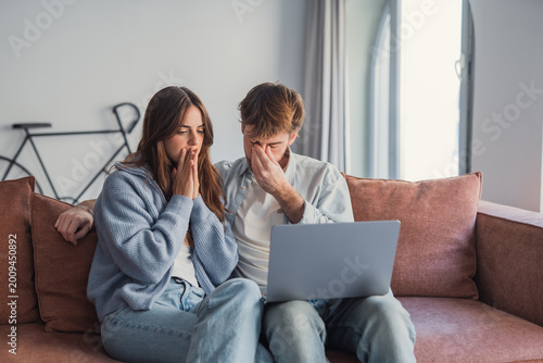 Cuadro en lienzo Young couple sits on a sofa looking shocked and worried while viewing something upsetting on a laptop, conveying stress, anxiety, relationship tension and financial or personal bad news at home