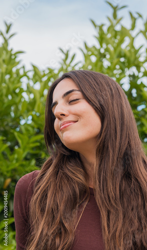 Принти на полотні A young woman with long brown hair smiles with closed eyes in a relaxed outdoor portrait amid lush green foliage, conveying calm, contentment, serenity and natural beauty