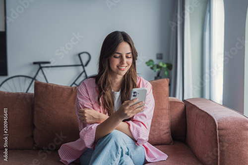Foto Young woman sits comfortably on a sofa in a bright living room, smiling as she uses her smartphone