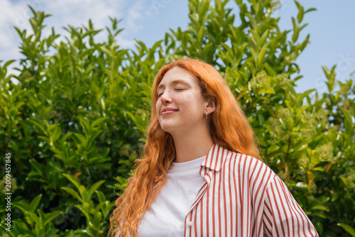 Obraz na płótnie A young redhead woman stands outdoors with eyes closed, breathing deeply and smiling softly amid lush green foliage, conveying calm, contentment and a relaxed summer vibe in casual striped shirt