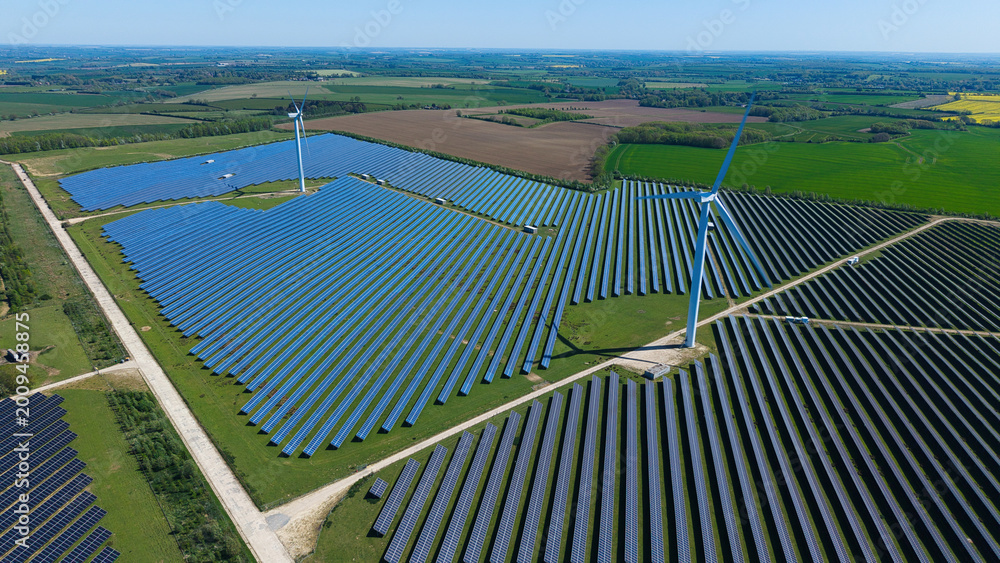 © AmazingAerialAgency - Aerial view of Chelveston Wind Farm featuring vast solar panel arrays and wind turbines across the green countryside in Wellingborough, England, United Kingdom.