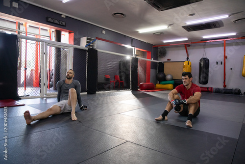 Fotografia Diverse male training partners sitting on mats at MMA gym cage, wearing rashguar