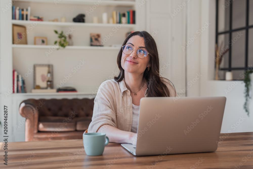 Fototapeta premium Young woman sits at a wooden desk in a cozy living-room home office, working on a laptop and smiling thoughtfully with a coffee mug nearby, conveying relaxed remote work, creativity and focus.