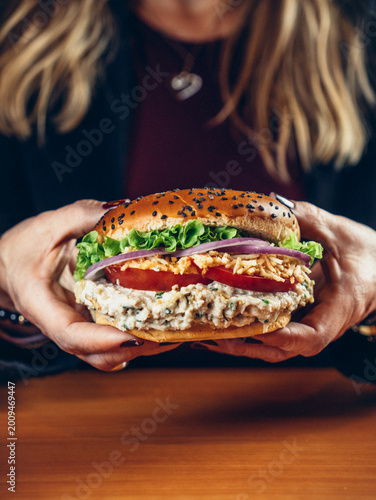 Chicken salad burger with lettuce, tomato, red onion, and crispy topping on a sesame bun, held in hands over a wooden table.
