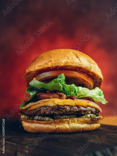 Juicy cheeseburger with bacon, lettuce, tomato, and pickles on a toasted bun, served on a dark surface with a vibrant red background.
