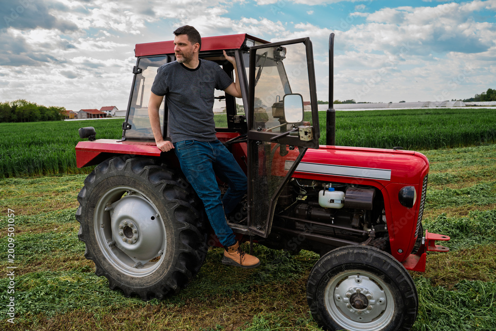 Obraz premium Handsome man sitting on the tractor and looking across the field, wheat in the background, agricultural farm