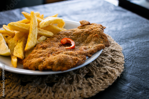 Detalle macro de una milanesa crujiente con enfoque selectivo en la textura del empanado, acompañada de papas fritas y una rodaja de limón fresco.