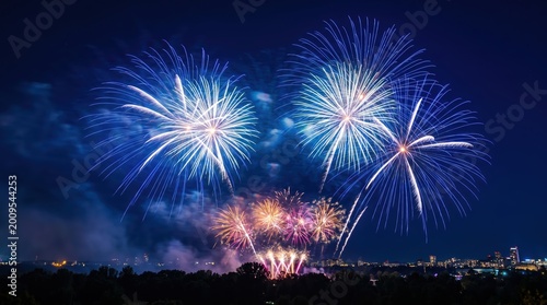 Bright blue and gold fireworks exploding in the night sky over a dark city skyline silhouette with light smoke trails and glowing sparks against a deep navy blue background during a celebration