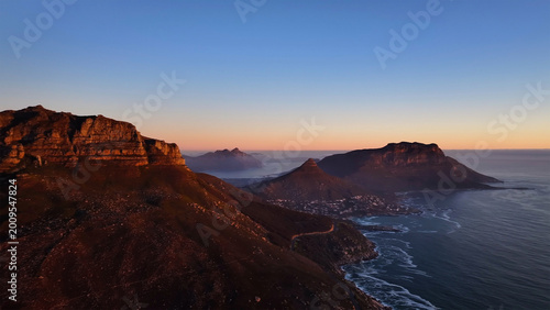 Aerial view of the rugged peaks of Table Mountain and Lion's Head overlooking the Atlantic coastline during a golden sunset in Cape Town, Western Cape, South Africa.