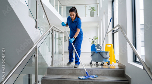 A professional cleaner mops a wet stair landing beside a cleaning cart in a bright modern office or apartment building.