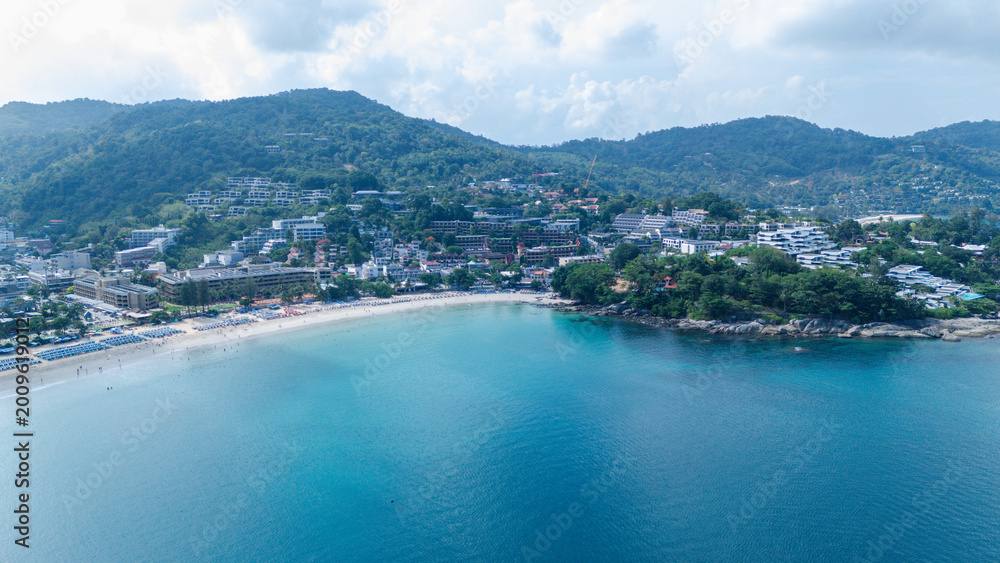 Obraz premium A coastal town nestled between green hills with a sandy beach meeting calm blue waters under a partly cloudy sky.