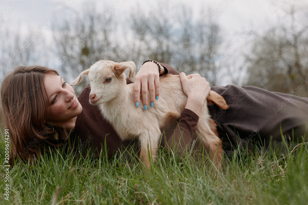 Fototapeta premium Young woman lying in grass outdoors, gently embracing a playful goat kid, capturing a warm moment of companionship, nature connection, soft light, relaxed mood, casual lifestyle.