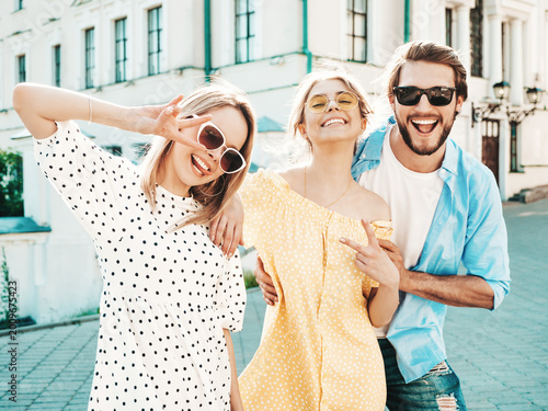 Group of young three stylish friends posing in the street. Fashion man and two cute dressed in casual summer clothes. Smiling models having fun in sunglasses.Cheerful women and guy at susnet