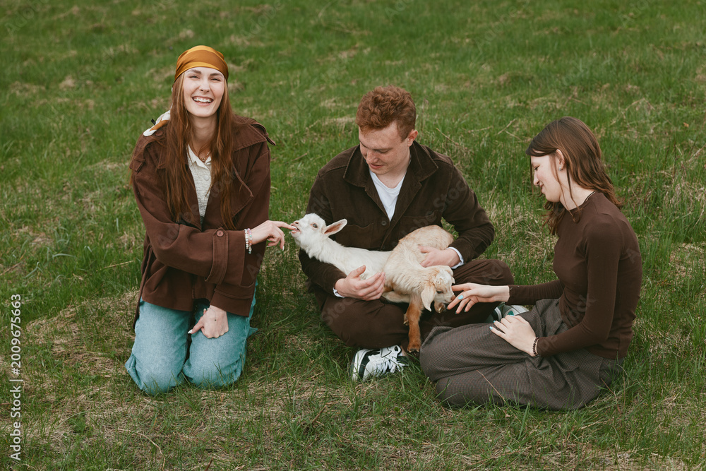 Obraz premium A warm outdoor scene features three friends kneeling on grass, gently petting a curious goat kid, capturing a lifestyle moment of friendship, care, and playful bond with animals in nature.