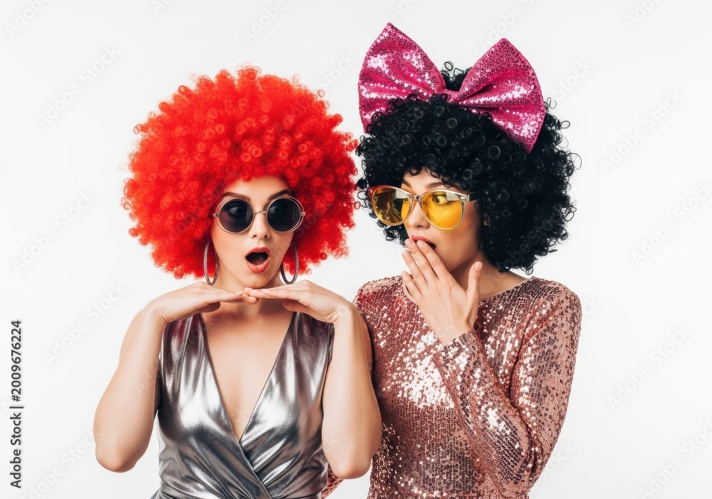 Fototapeta premium Two surprised young women in colorful afro wigs and sequined outfits pose against a white background.