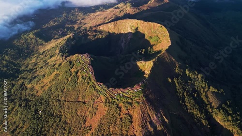 Volcanic terrain with deep caldera and steep slopes under golden light highlighting rugged mountain and surrounding forested landscape in wide aerial drone view