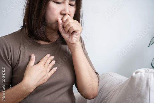 Asian woman feeling nauseous and coughing, holding her chest and covering mouth at home. Concept of acid reflux, food poisoning, morning sickness, or flu. Female suffering from illness or GERD