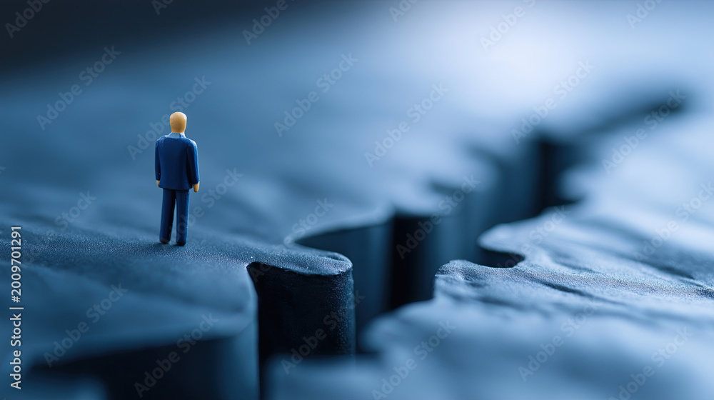 © sam richter - A lone miniature business figure stands precisely on the edge of a vast, deep blue canyon crack, representing concepts of risk and decision.