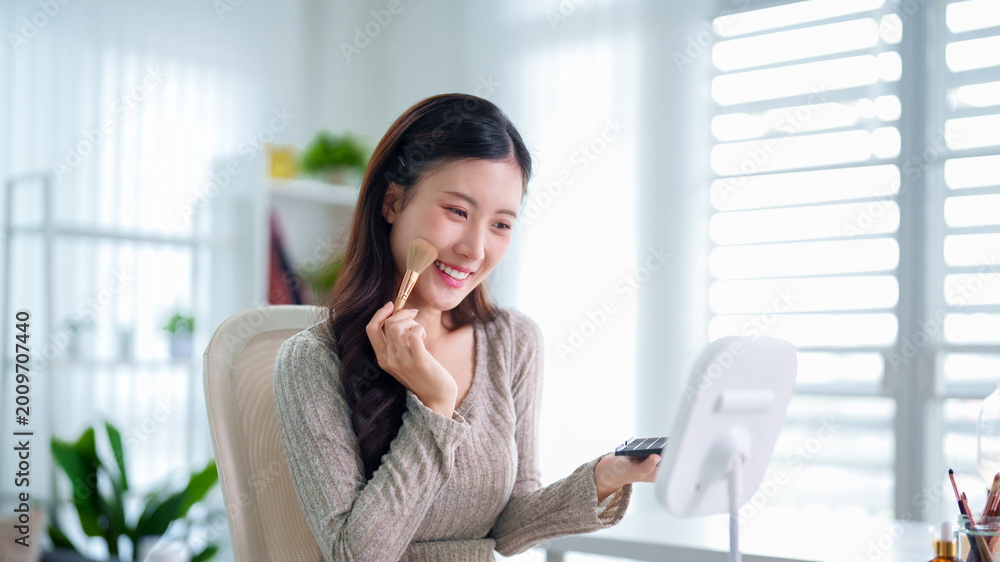 Fototapeta premium beautiful young Asian women applying makeup with cosmetic brush sitting home office modern interior bright natural light enjoying beauty care routine lifestyle