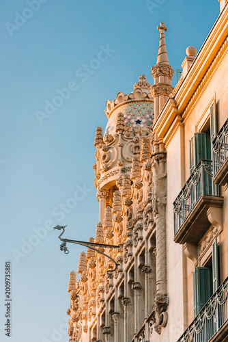 Detailed stone facade of a historic Modernisme building in Barcelona, featuring a decorative dome and ornate sculptures against a blue sky.