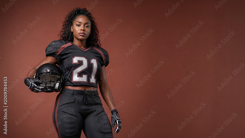 Obraz premium Confident African American female tackle football player in a black uniform holds a helmet against a warm brown studio background with ample copy space.
