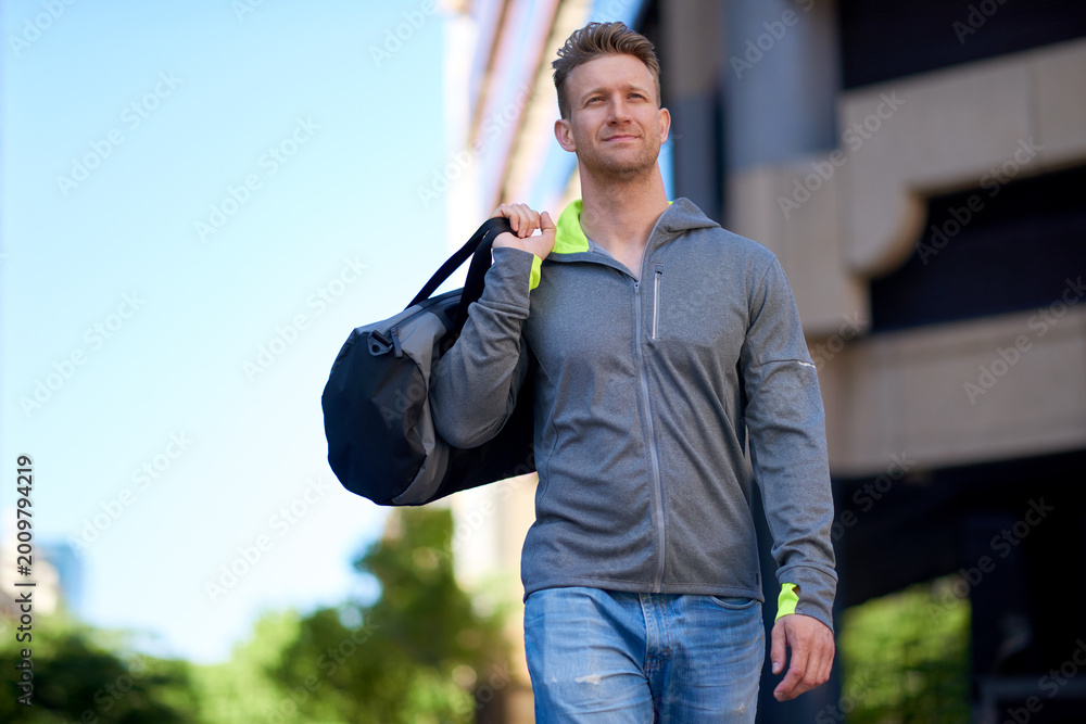 Fototapeta premium Man, fitness and walking with gym bag in city for workout equipment or outdoor preparation. Active, male person or athlete getting ready with exercise gear for health and wellness in an urban town