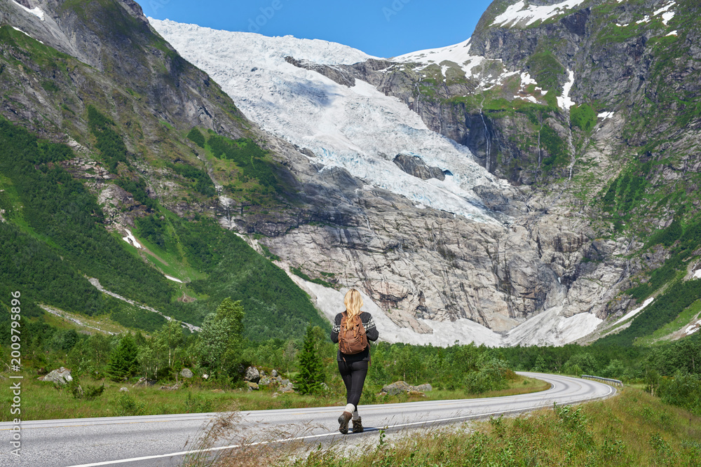 Fototapeta premium Back, woman and hiking on road with backpack for travel, adventure and backpacking holiday. Mountain, female person and walking for trekking journey, glacier scenery and explore nature on vacation
