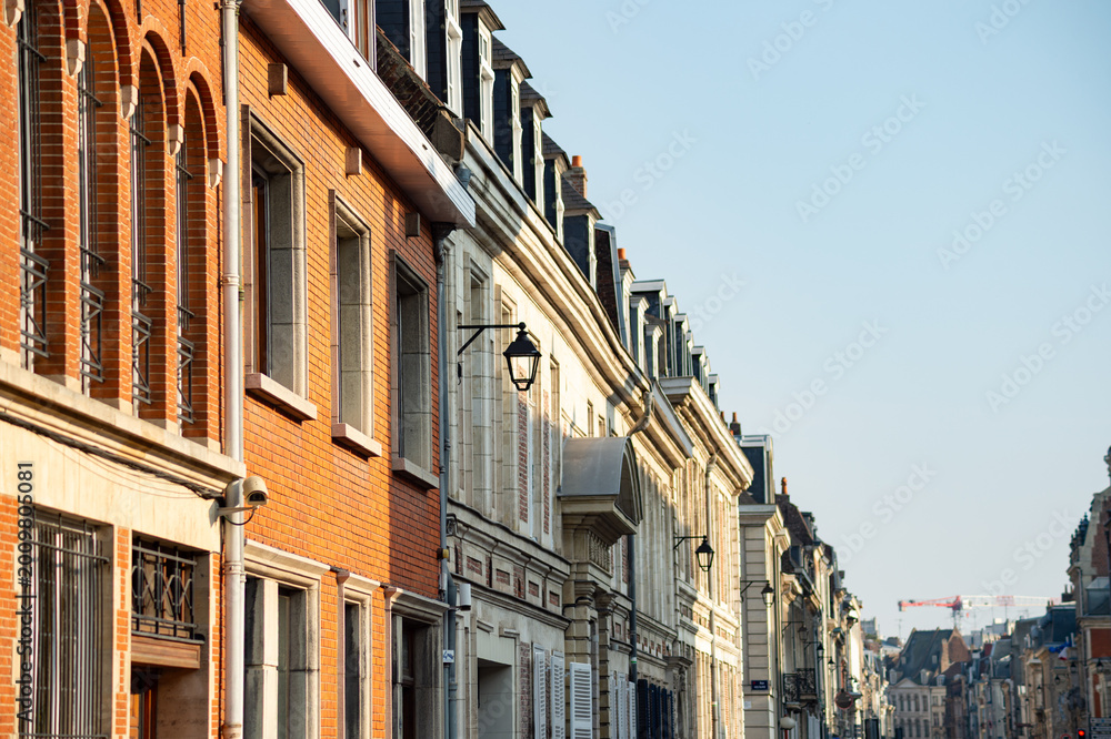 Fototapeta premium Row of historic brick and stone buildings on a street in Lille, France. Architecture features arched windows, dormers, and traditional street lanterns under a clear sky.