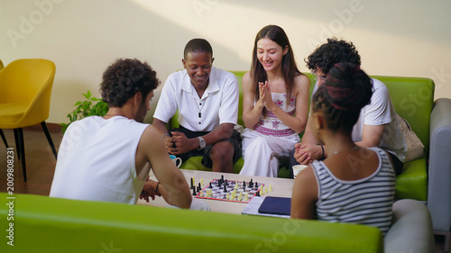 Indian young adult gen z male female group team sitting on sofa couch play chess indoor room place teen girl boy look game watch match talk speak share idea spend break time enjoy new day have fun joy