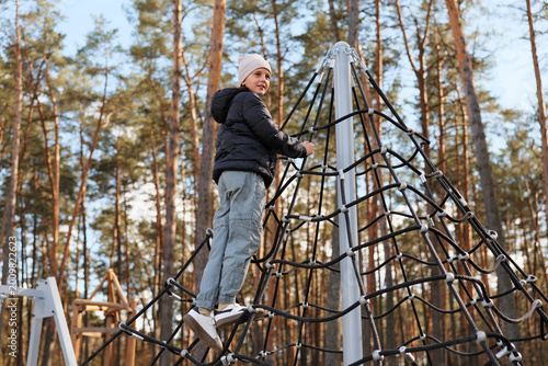A child is climbing a netted structure at a playground located in a forest. The trees surround the play area under a clear blue sky