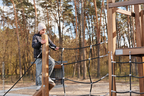 A child climbs on a wooden structure in a park surrounded by tall trees. The sun shines as the child enjoys the activity in the open space