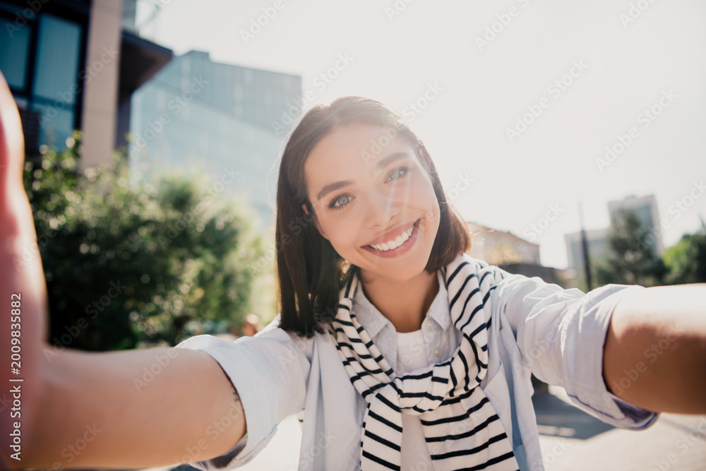 Fototapeta premium Young woman smiling and taking a selfie outdoors in sunny urban street, casual striped scarf, natural light portrait capturing happy lifestyle and travel vibes