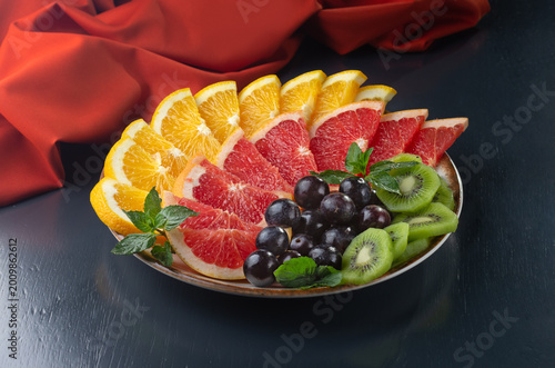 plate of fruit with oranges, grapefruit, kiwis, and grapes. The plate is white and the fruit is colorful on a dark background