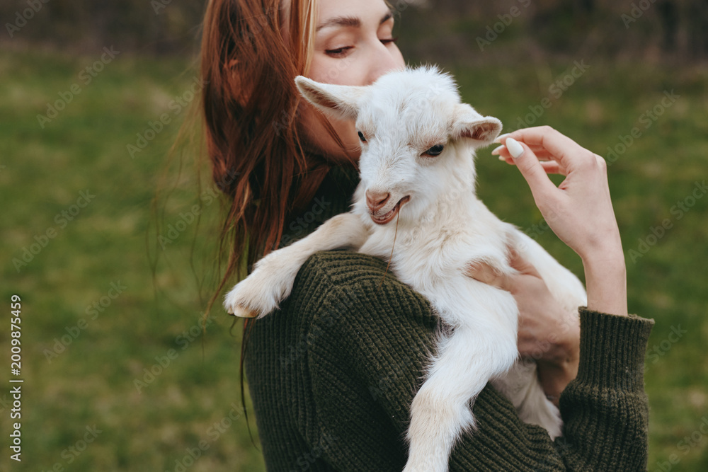 Fototapeta premium Goat, kid, woman outdoors, tender moment as she holds a gentle goat kid, sunlit grass, casual sweater, natural colors, candid lifestyle scene, outdoor companionship