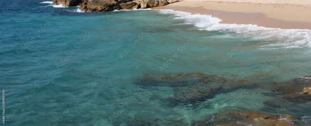 Obraz premium Clear turquoise ocean waves wash onto a sandy beach. Rocky formations border the water and shore. Sunlit water reveals underwater rocks and sand.
