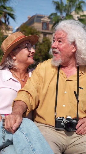 Cheerful elderly couple sitting on a park bench, laughing and talking. Happy senior man and woman enjoying a sunny day on vacation