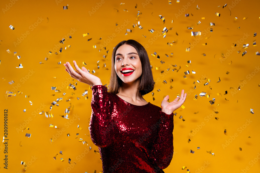 Fototapeta premium Happy woman celebrating with confetti in red sequin dress on yellow background, festive party portrait expressing joy and glamour