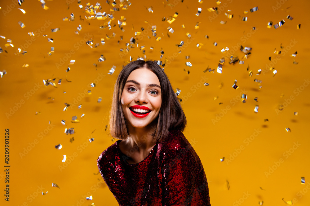 Naklejka premium Smiling woman in red sequin dress celebrating with silver confetti against a vibrant yellow background, joyful glamorous party portrait
