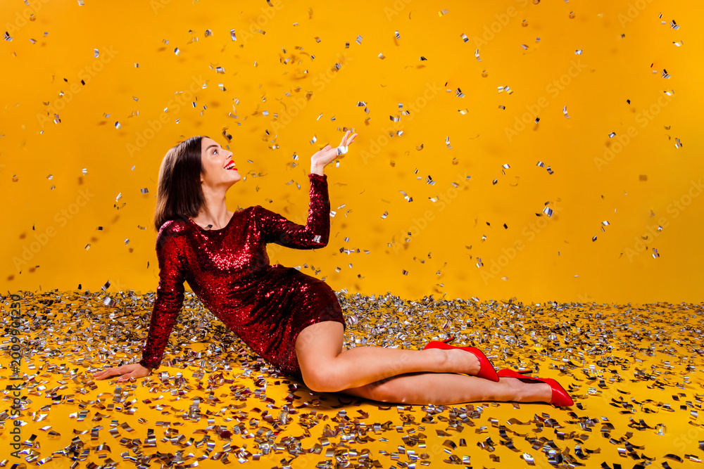Obraz premium Woman in red sequin dress lying among confetti on yellow background celebrating a festive party with glamorous heels and joyful expression